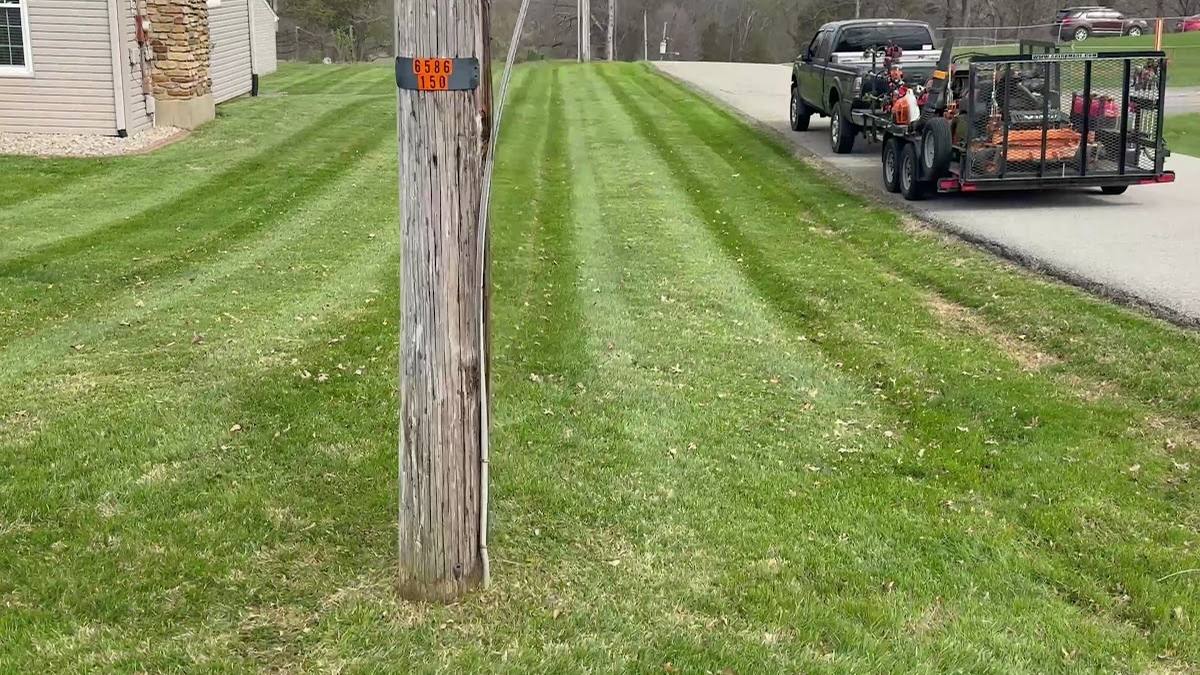 A freshly cut front lawn with clean mowing stripes running to the street, with the crew's truck and loaded equipment trailer parked at the curb.